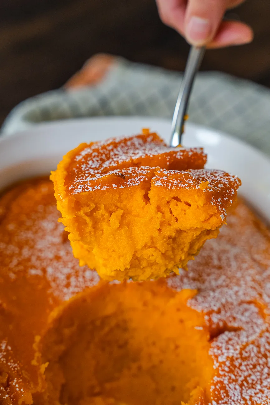 A metal spoon lifting a tender portion of baked carrot souffle from a white baking dish.
