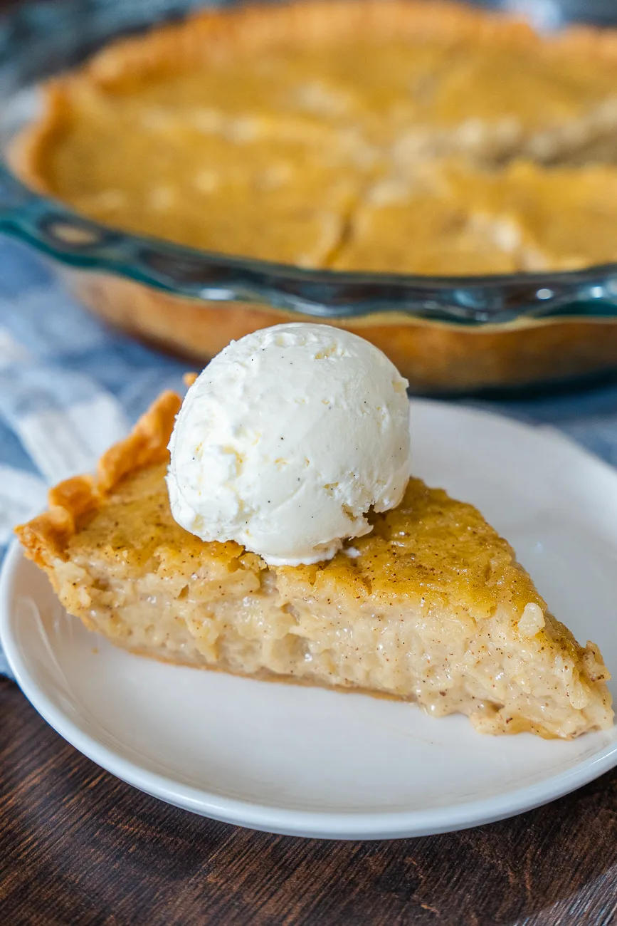 A single slice of Marlborough pie on a white dessert plate topped with a scoop of vanilla ice cream, beside a whole pie in a clear baking dish.