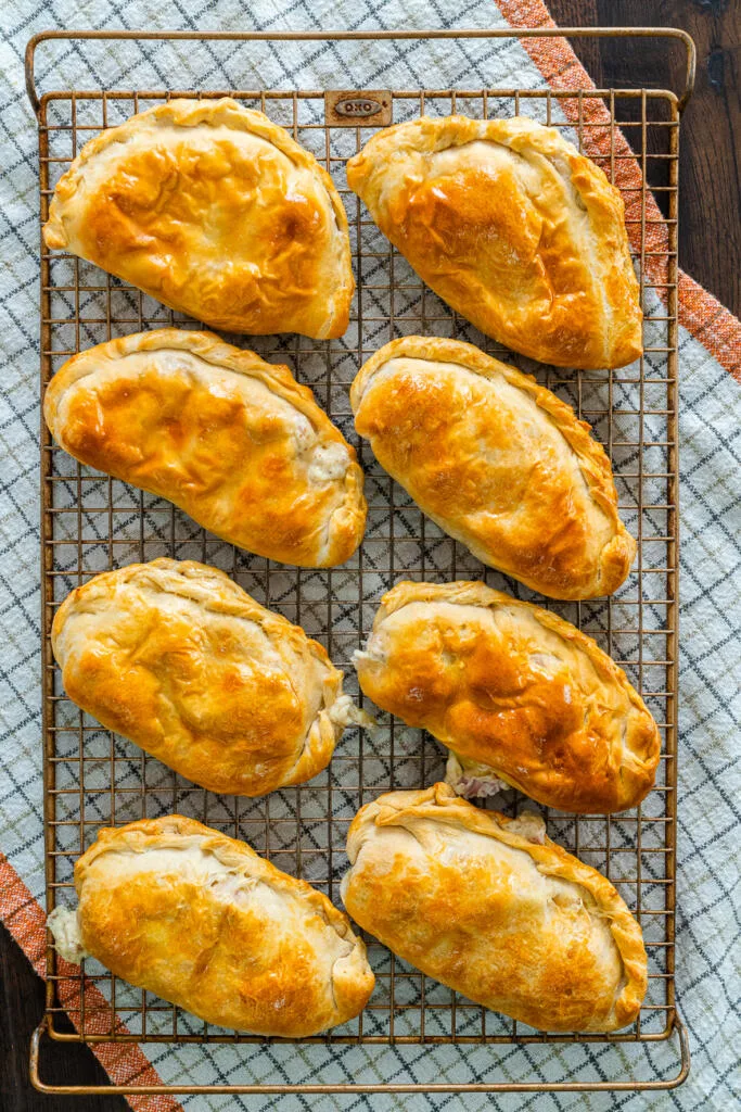 Eight golden brown baked hand pies cooling on a metal wire rack over a checkered cloth.