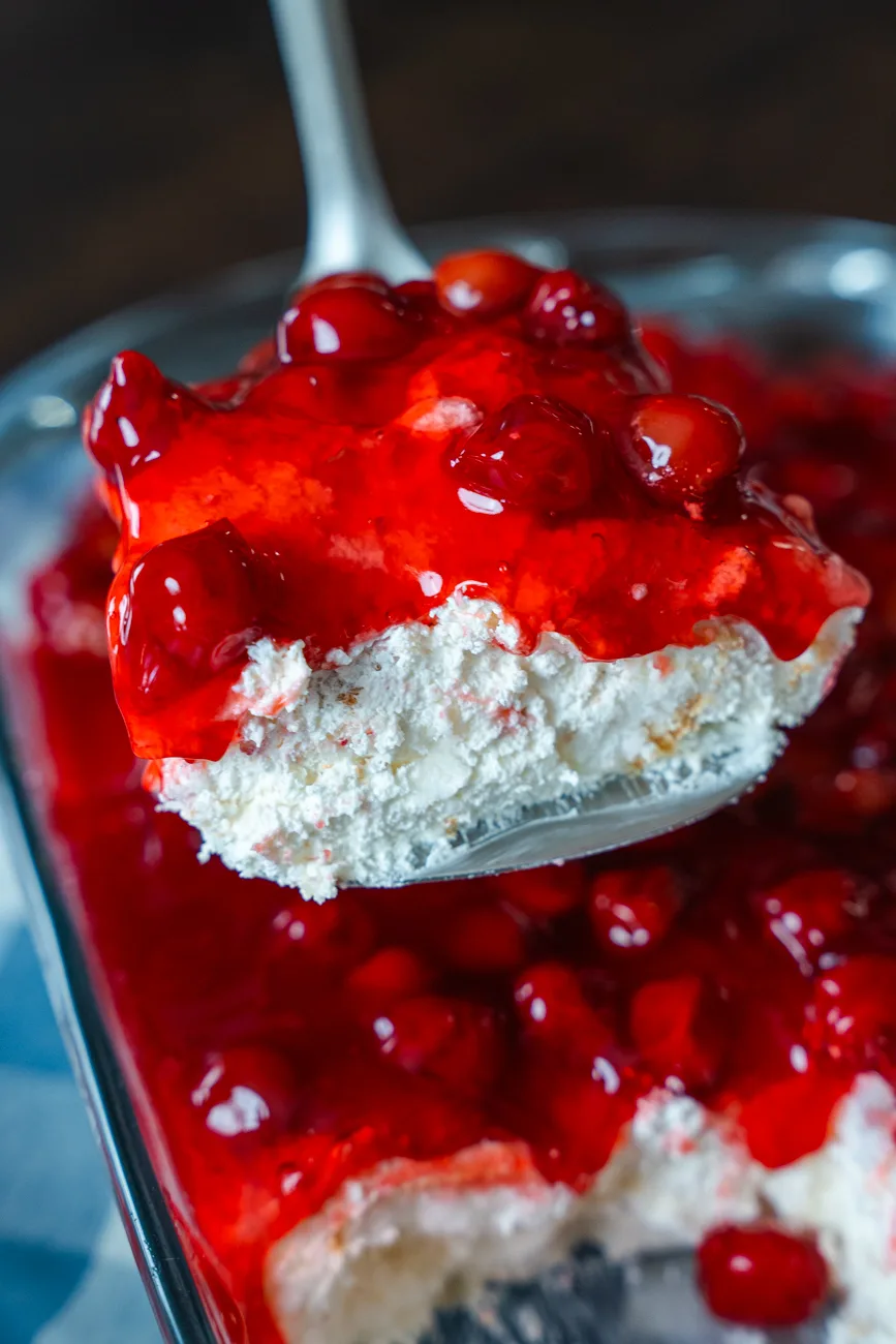Close-up of a metal spoon lifting a serving of Cherries in the Snow showing glossy cherry pie filling over a fluffy white cake mixture.