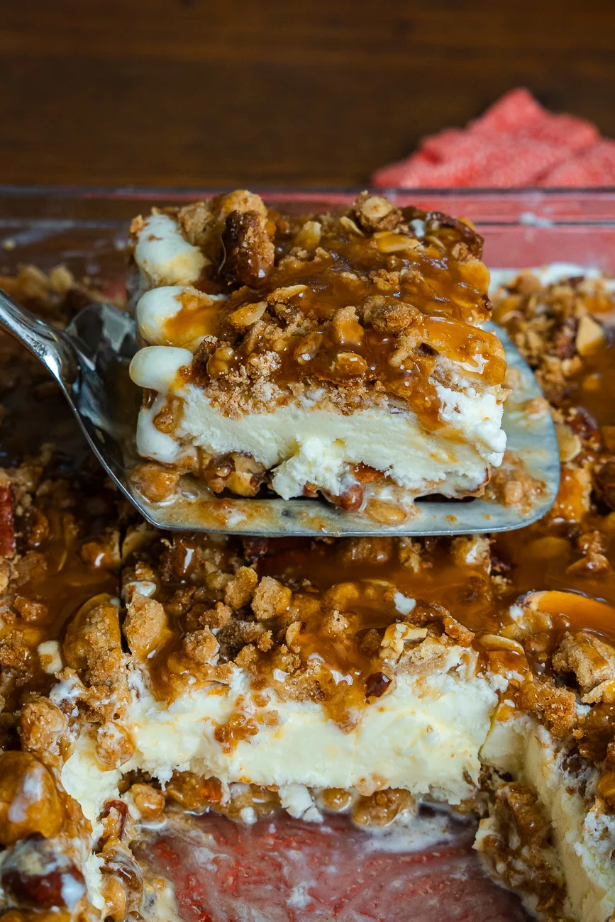 A metal spatula lifting a neat square of frozen vanilla ice cream dessert layered with oat-nut crumble and butterscotch from a glass baking pan.